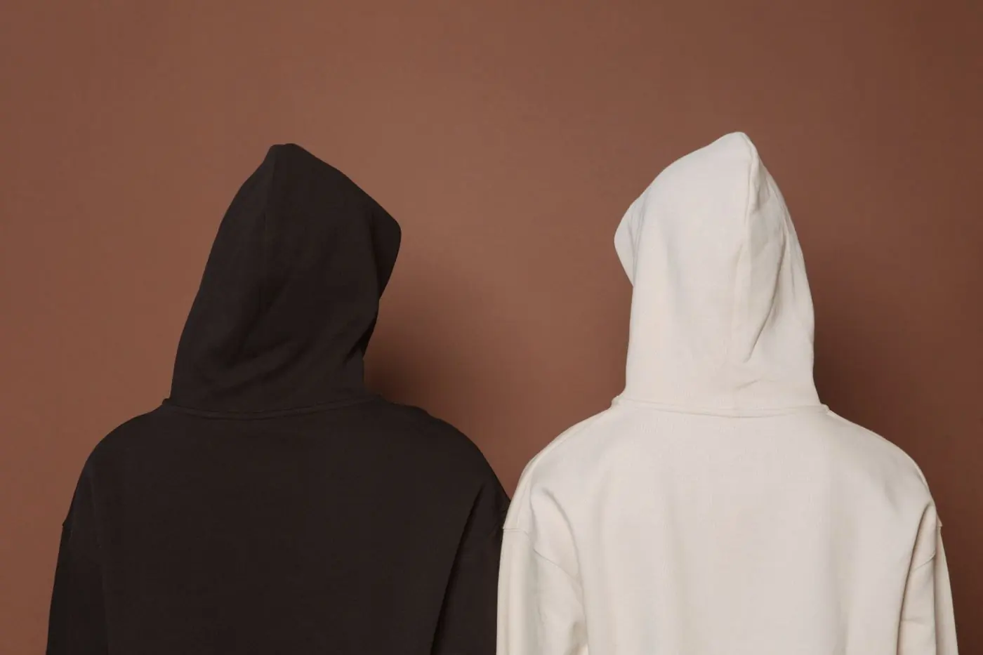 Two plain hoodies from behind, one black and one cream, against a brown wall